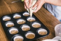 From above crop female confectioner standing at table with muffin tray and filling paper liners with batter in kitchen at home — Stock Photo