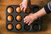 Blick von oben auf anonyme Senior-Koch nimmt köstliche Muffin in Pappschachtel aus Tablett auf Holztisch in der Küche platziert — Stockfoto