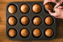 Vue du dessus du cuisinier anonyme prenant un délicieux muffin dans une caisse en papier à partir d'un plateau placé sur une table en bois dans la cuisine — Photo de stock