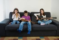 Menino e meninas positivos em roupas casuais sentados no sofá confortável juntos e tocando instrumentos de música enquanto passam o tempo juntos em casa — Fotografia de Stock