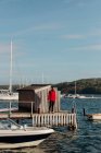 Мужской турист в красной теплой куртке, стоящий в одиночестве на деревянном пирсе с лодками и яхтами в солнечный день в выходные и смотрящий в сторону — стоковое фото