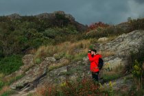 Vue latérale du touriste masculin en veste rouge et avec sac à dos debout sur une colline rocheuse et prenant des photos du paysage par temps nuageux — Photo de stock