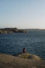 Vue arrière de touriste féminine anonyme en vêtements de dessus chauds décontractés et avec sac à dos assis sur le bord de la mer rocheuse et admirant une vue imprenable sur la mer par temps venteux — Photo de stock