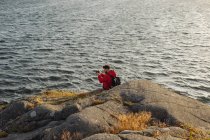 Turista masculino em jaqueta vermelha casual e com mochila sentada em penhasco rochoso e tirando fotos da bela paisagem marinha — Fotografia de Stock