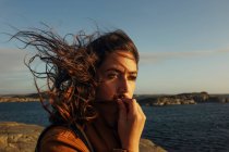 Красивая молодая женщина-путешественница в повседневной одежде стоит на скалистом берегу моря с волосами, дующими на ветру и мечтательно глядя в сторону — стоковое фото