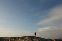Вид сбоку на туриста в красной куртке и с рюкзаком прогуливаясь по скалистой склоне холма держа фотокамеру и фотографируя красивый пейзаж в пасмурный день — стоковое фото