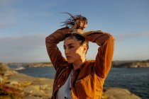 Beautiful young female traveler in casual clothes standing on rocky seashore holding hair up with hand in a high ponytail during a windy day and looking away dreamily — Stock Photo