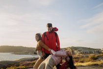Eine Gruppe gut gelaunter junger Touristen in lässiger Oberbekleidung und mit Rucksäcken und Fotokamera steht zusammen und hält Händchen an felsigen Klippen am sonnigen Meeresufer — Stockfoto