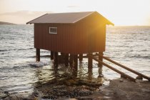 Casa flotante marrón con pequeña ventana y puerta instalada en pontón cerca de la costa del mar con olas tocando la superficie de la costa al amanecer - foto de stock