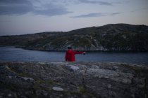 Rückansicht eines entlegenen Touristen in rotem lässigem Kapuzenpulli, der auf einer felsigen Klippe in der Nähe des Meeres steht und mit dem Finger zur Seite zeigt, während er die Kamera in der Hand hält — Stockfoto