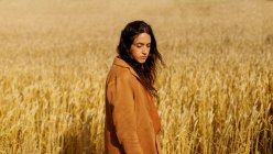 Menina pensativa em casaco elegante e cabelo encaracolado marrom em pé no meio do campo de centeio de ouro sem fim e olhando para baixo — Fotografia de Stock