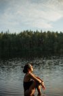 Desde arriba vista lateral de hembra delgada en traje de baño sentado con los ojos cerrados en aguas tranquilas del lago mientras disfruta de la puesta del sol y el paisaje majestuoso - foto de stock