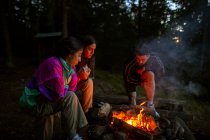 Compañía de gente amigable en ropa casual que se reúne alrededor de la hoguera en madera mientras hace fuego y se calienta por la noche - foto de stock