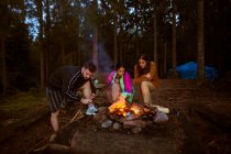 Company of friendly people in casual clothes gathering around bonfire in wood while making fire and warming up in evening — Stock Photo