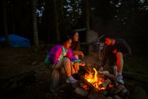 Компания дружелюбных людей в повседневной одежде собирается вокруг костра в лесу, разжигая огонь и согреваясь вечером — стоковое фото