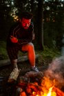Спокойный мужчина-кемпер в повседневной одежде стоит с бревном возле костра ночью и согревается во время кемпинга в лесу — стоковое фото