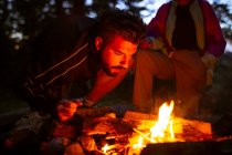 Calma campista masculino en ropa casual de pie con tronco cerca de la hoguera por la noche y el calentamiento durante el campamento en madera - foto de stock