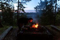 Calma campista masculino en ropa casual de pie con tronco cerca de la hoguera al atardecer y calentamiento durante el campamento en madera - foto de stock