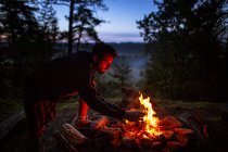 Спокойный мужчина-кемпер в повседневной одежде стоит с бревном возле костра на закате и согревается во время кемпинга в лесу — стоковое фото