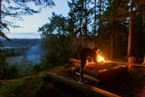 Спокойный мужчина-кемпер в повседневной одежде стоит с бревном возле костра на закате и согревается во время кемпинга в лесу — стоковое фото