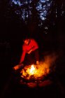 Calma campista masculino en ropa casual de pie con tronco cerca de la hoguera por la noche y el calentamiento durante el campamento en madera - foto de stock
