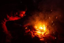 Calmo macho campista em casual desgaste de pé com log perto de fogueira à noite e aquecimento durante o acampamento em madeira — Fotografia de Stock
