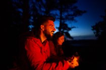 Junges Paar zeltet zusammen in Freizeitkleidung mit Baumstamm am Lagerfeuer bei Sonnenuntergang und wärmt sich beim Zelten im Wald auf — Stockfoto