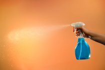 Persona anonima con spray in bottiglia di plastica per la disinfezione delle superfici polverizzando liquido su fondo arancione in studio — Foto stock
