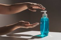Crop faceless person pulling hands for plastic blue liquid soap bottle with white dispenser inside located on white table with shadow — Stock Photo