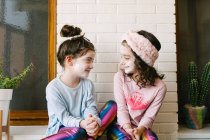 Fröhlich lachende Mädchen in ähnlichen Kleidern und aufgesetzter Gesichtsmaske sitzen zu Hause auf einem Holztisch und haben Spaß vor ziegelsteinweißer Wand — Stockfoto