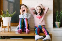 Радостные смеющиеся девушки в похожей одежде и в маске для лица сидят дома на деревянном столе и веселятся у кирпичной белой стены — стоковое фото