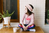 Счастливая улыбающаяся маленькая девочка с вьющимися волосами в розовой пижаме и повязкой на голове, сидящей возле белой кирпичной стены в маске для лица — стоковое фото
