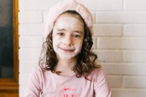 Feliz sorrindo criança pequena fêmea com cabelos cacheados em pijama rosa e headband sentado perto da parede de tijolo branco na máscara facial e olhando para a câmera — Fotografia de Stock