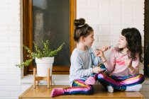 Hermanitas sonrientes sentadas en la mesa y comiendo deliciosos dulces azules masticables divirtiéndose y mirándose mientras descansan en casa durante el fin de semana - foto de stock
