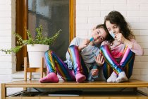 Улыбающиеся сёстры сидят на столе и едят вкусные голубые жевательные конфеты, развлекаясь и глядя друг на друга, отдыхая дома в выходные. — стоковое фото