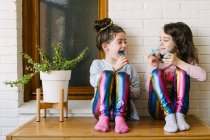 Усміхнені маленькі сестри сидять на столі і їдять смачні блакитні жувальні цукерки, розважаючись і дивлячись один на одного, відпочиваючи вдома у вихідні — стокове фото