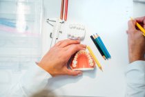 Сверху растительного ортодонта с карандашом в руке, работающим с белой зубной плесенью за столом с профессиональным оборудованием — стоковое фото