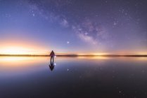 Silhueta de homem anônimo em pé na superfície de reflexão da água e estendendo a mão para o céu estrelado colorido noite com forma leitosa — Fotografia de Stock