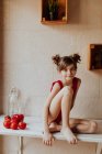 Carino ragazza a piedi nudi in body rosso seduto sul bancone con pomodori e pepe rosso in cucina — Foto stock