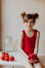 Cute barefoot girl in red bodysuit and with strawberries in her hair holding red pepper looking at camera sitting on counter near tomatoes in kitchen — Stock Photo