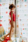 Vue latérale de la fille en body rouge debout sur un canapé doux et regardant par la fenêtre à travers des rideaux colorés à la maison — Photo de stock
