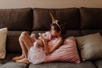 Vista lateral da menina descalça em traje de unicórnio abraçando brinquedo de pelúcia enquanto descansa no sofá confortável em casa — Fotografia de Stock