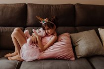 Seitenansicht des barfüßigen Mädchens im Einhornkostüm, das Plüschtier umarmt, während es sich zu Hause auf der bequemen Couch ausruht — Stockfoto