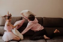 Вид збоку босоніж підліток хлопець вдарив сестру в однотонному костюмі з подушкою, граючи на дивані разом — стокове фото