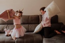 Вид збоку босоніж підліток хлопець вдарив сестру в однотонному костюмі з подушкою, граючи на дивані разом — стокове фото
