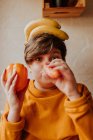 Chubby teen boy with bananas on head smiling and playing with pepper and carrot in kitchen — Stock Photo