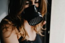 Несчастная молодая женщина в черной защитной маске, стоящая возле стеклянной стены в закрытой комнате, представляя концепцию ограничения и изоляции во время вспышки коронавируса — стоковое фото