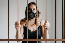Jeune femme en masque de protection respiratoire et robe noire debout derrière une clôture métallique et regardant la caméra tout en représentant le concept de prévention et d'isolement du coronavirus — Photo de stock