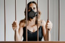 Young female in protective respirator mask and black dress standing behind metal fence and looking at camera while representing concept of coronavirus prevention and isolation — Stock Photo