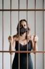 Молодая женщина в защитной респираторной маске и черном платье стоит за металлическим забором и смотрит в камеру, представляя концепцию коронавирусной профилактики и изоляции — стоковое фото